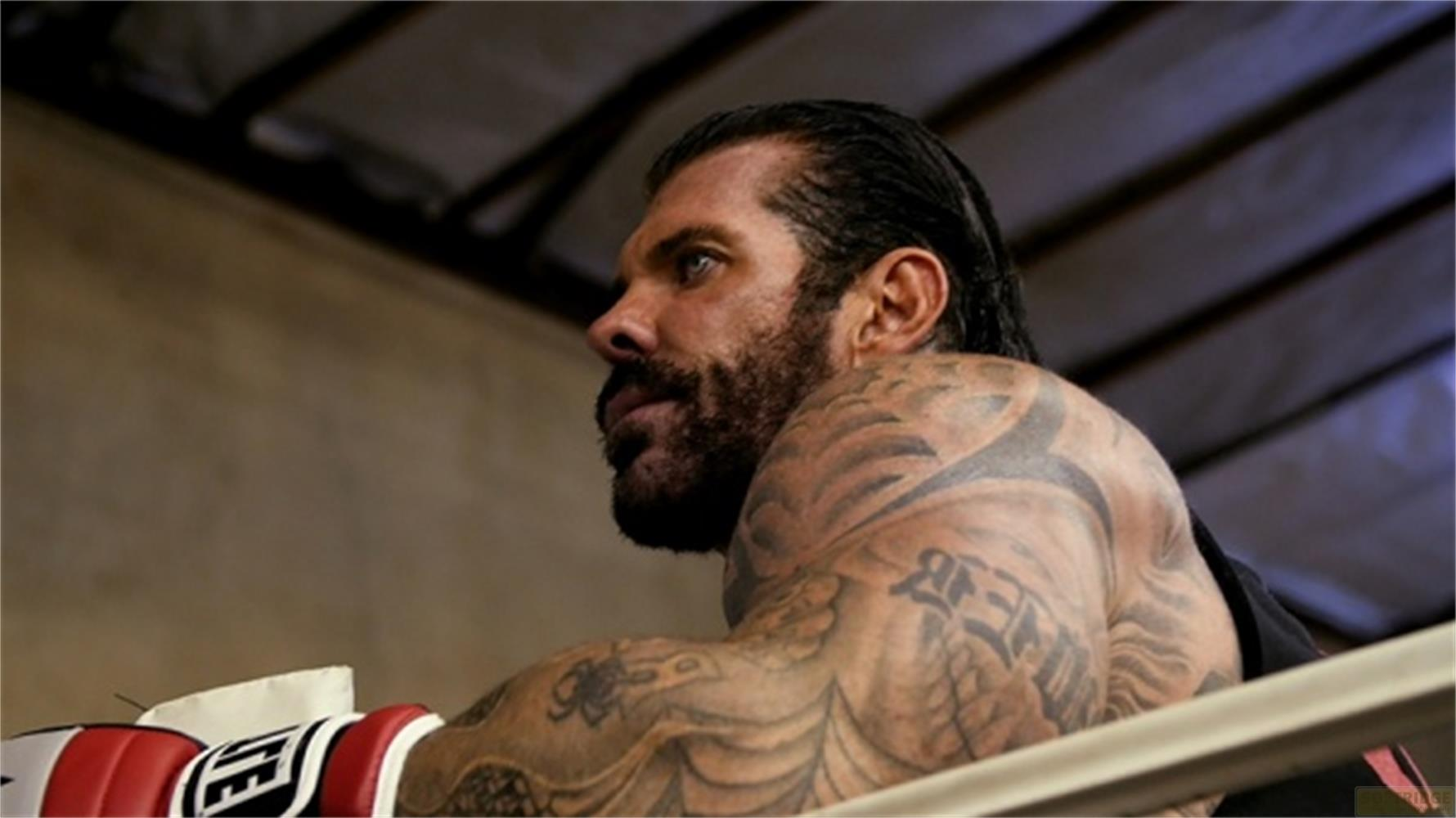 Un uomo con barba e tatuaggi in un ring di pugilato, indossa guanti rossi e bianchi.