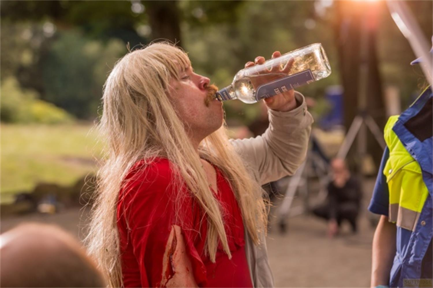 Persona con capelli biondi beve da una bottiglia in un parco.