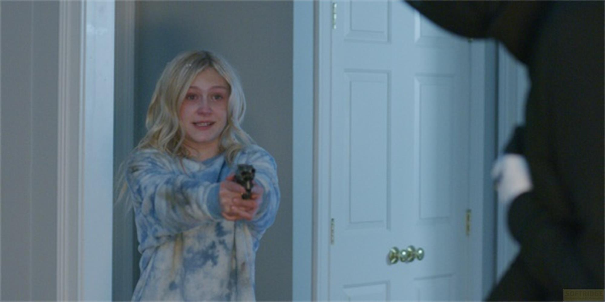 Una giovane ragazza bionda tiene una pistola mentre una persona in nero è in primo piano.