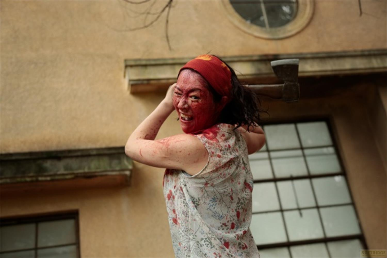 Una donna coperta di sangue con un'ascia la solleva per colpire davanti a un edificio.
