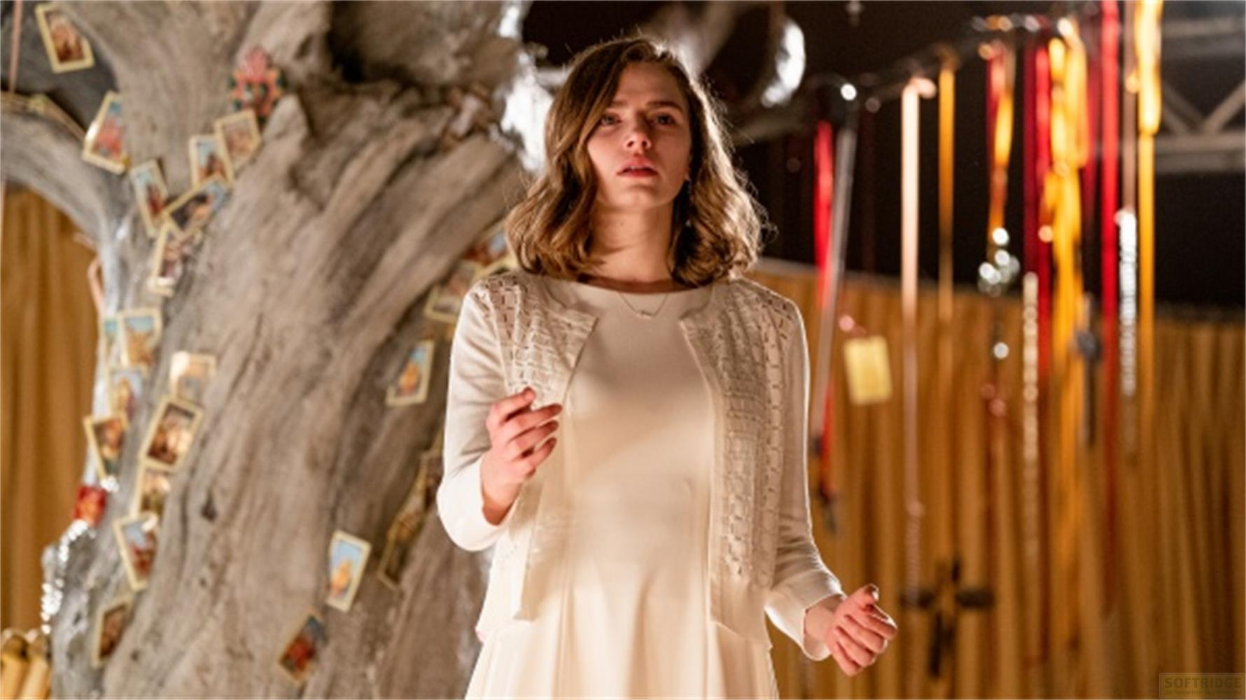 Una donna in abito bianco e cardigan sta davanti a un albero con carte e nastri colorati.