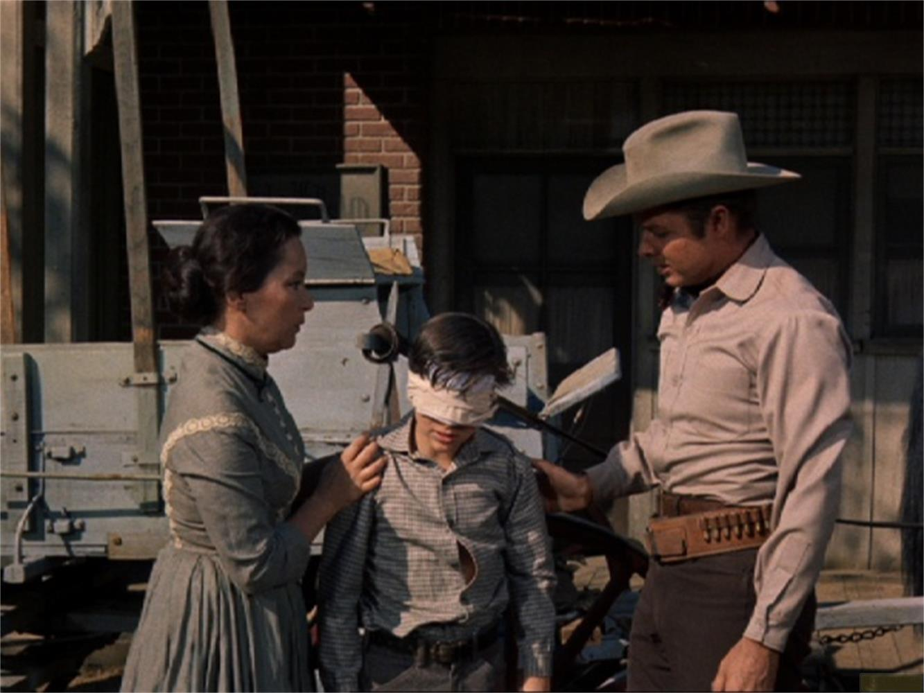 Uomo con cappello e donna bendano un ragazzo. All'aperto, stile western.