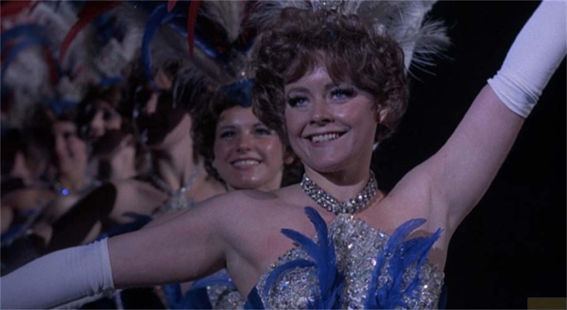 Une femme coiffée de plumes et vêtue de bleu sourit.