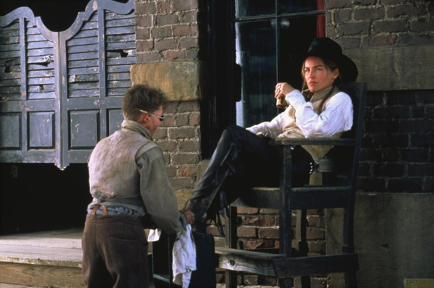 Un homme en tenue western est assis sur une chaise et boit. Un autre homme se penche.