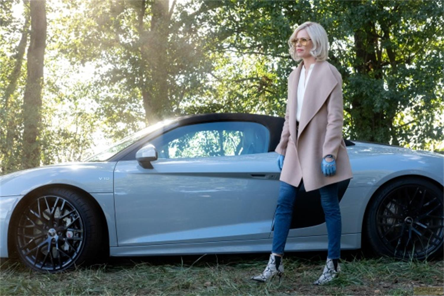 Una donna in jeans e cappotto sta accanto a una macchina sportiva argentata sull'erba.