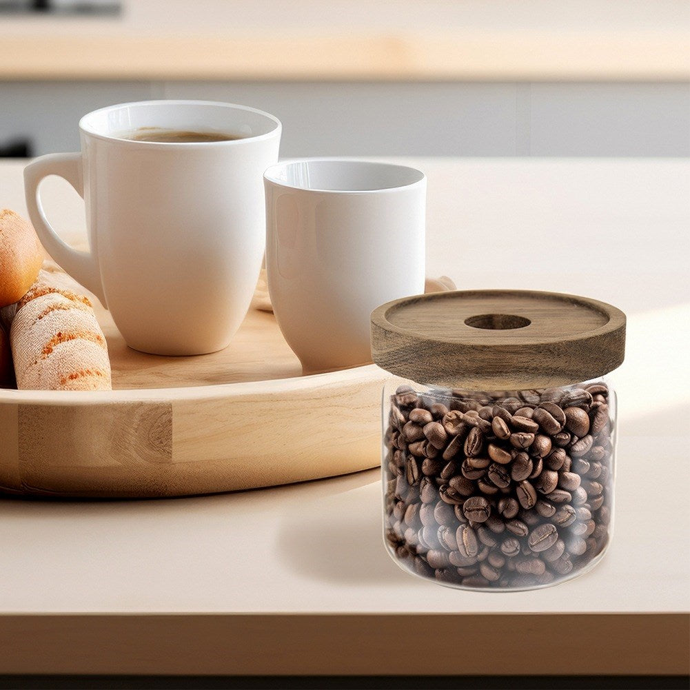Granos de café en un frasco de vidrio con tapa de madera, junto a dos tazas blancas y pasteles.