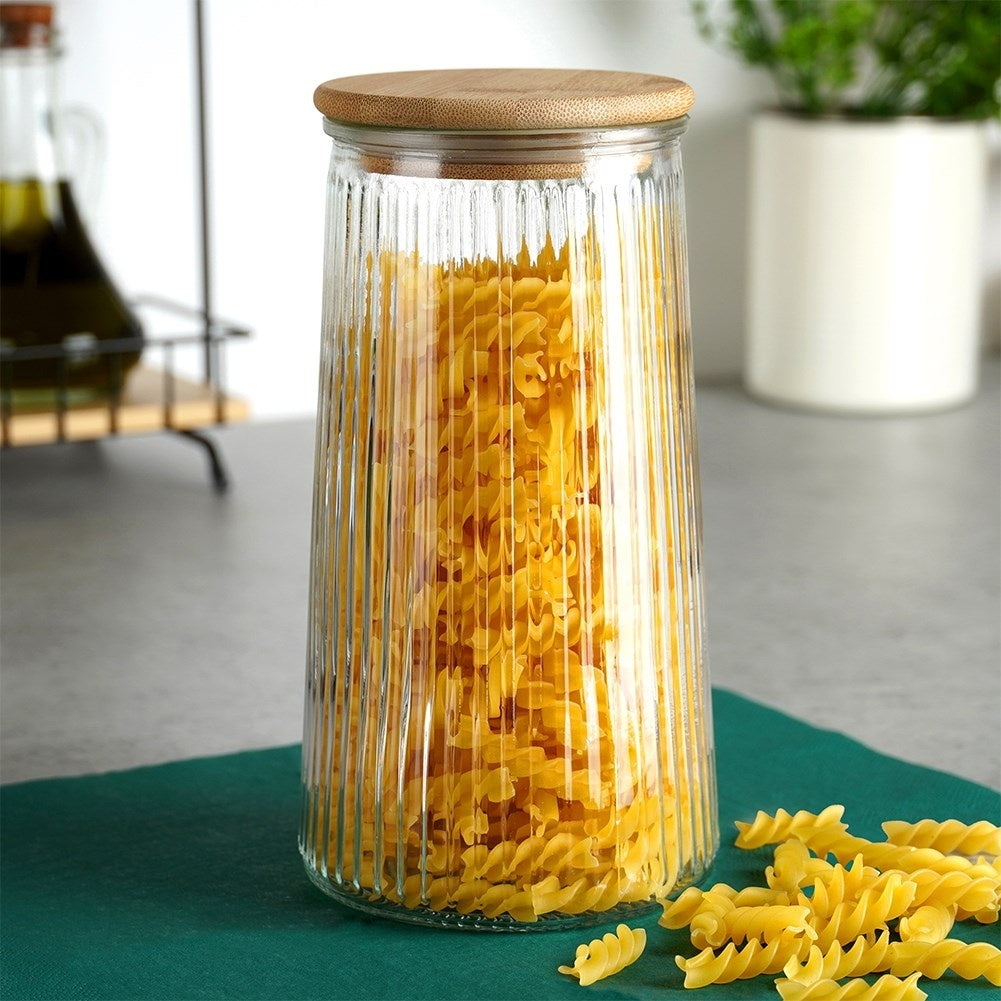 Un barattolo di vetro trasparente e scanalato con coperchio in legno, pieno di pasta fusilli, poggia su un tappetino verde scuro. Altra pasta si rovescia sul tappetino.