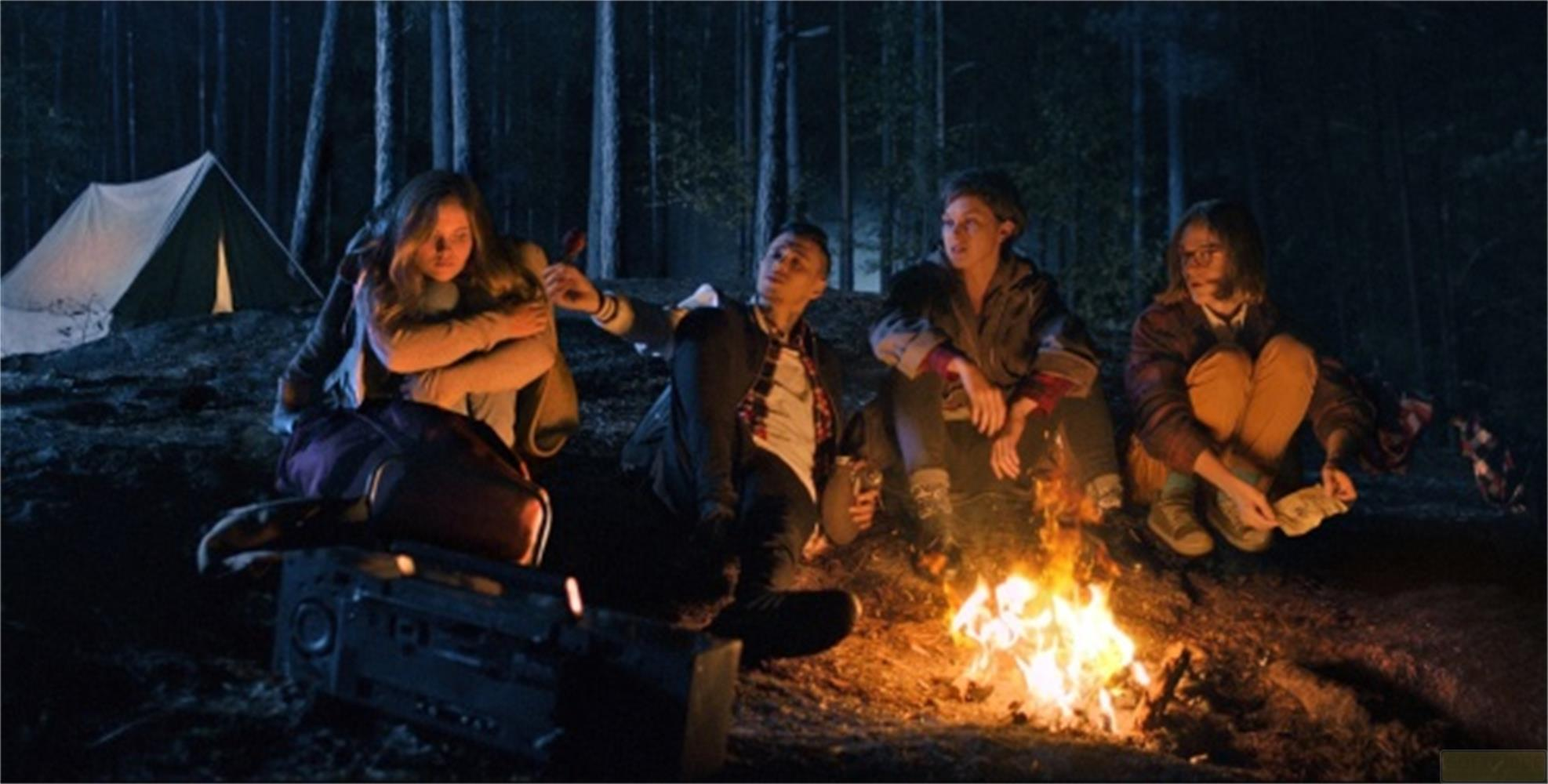 Quattro amici seduti attorno a un falò di notte in una foresta. Una tenda bianca sullo sfondo.