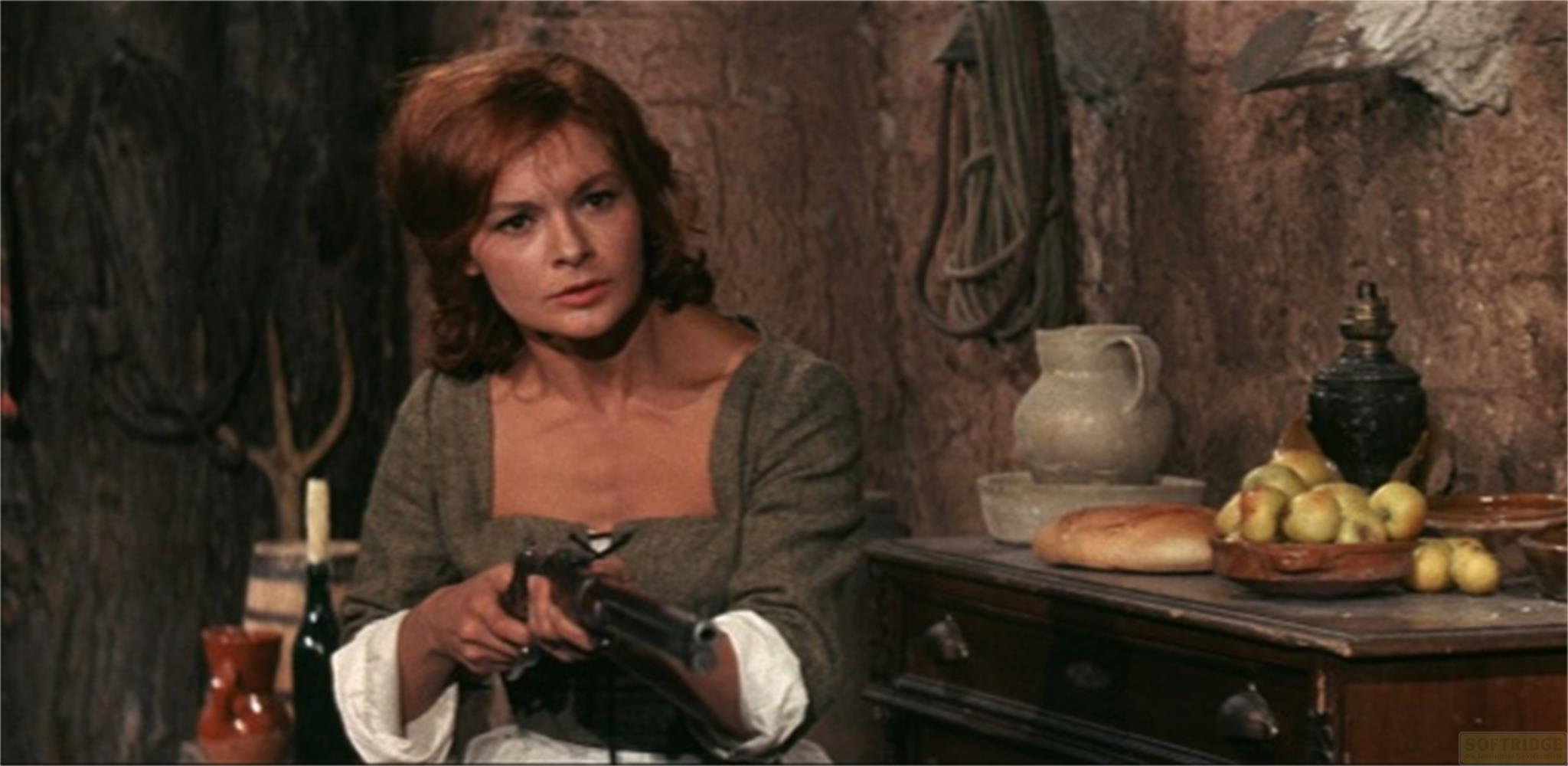 Donna con fucile in ambiente rustico, con frutta e pane su una cassettiera.