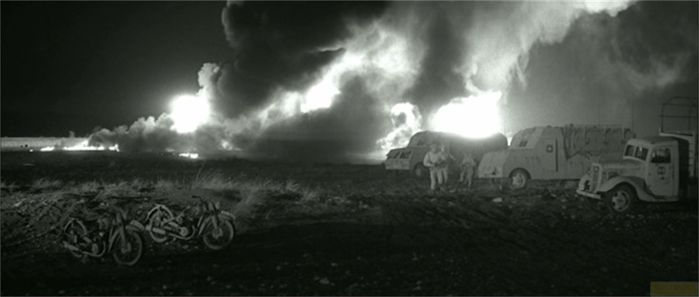 Veicoli militari e motociclette parcheggiati vicino a un campo con colonne di fumo di notte.