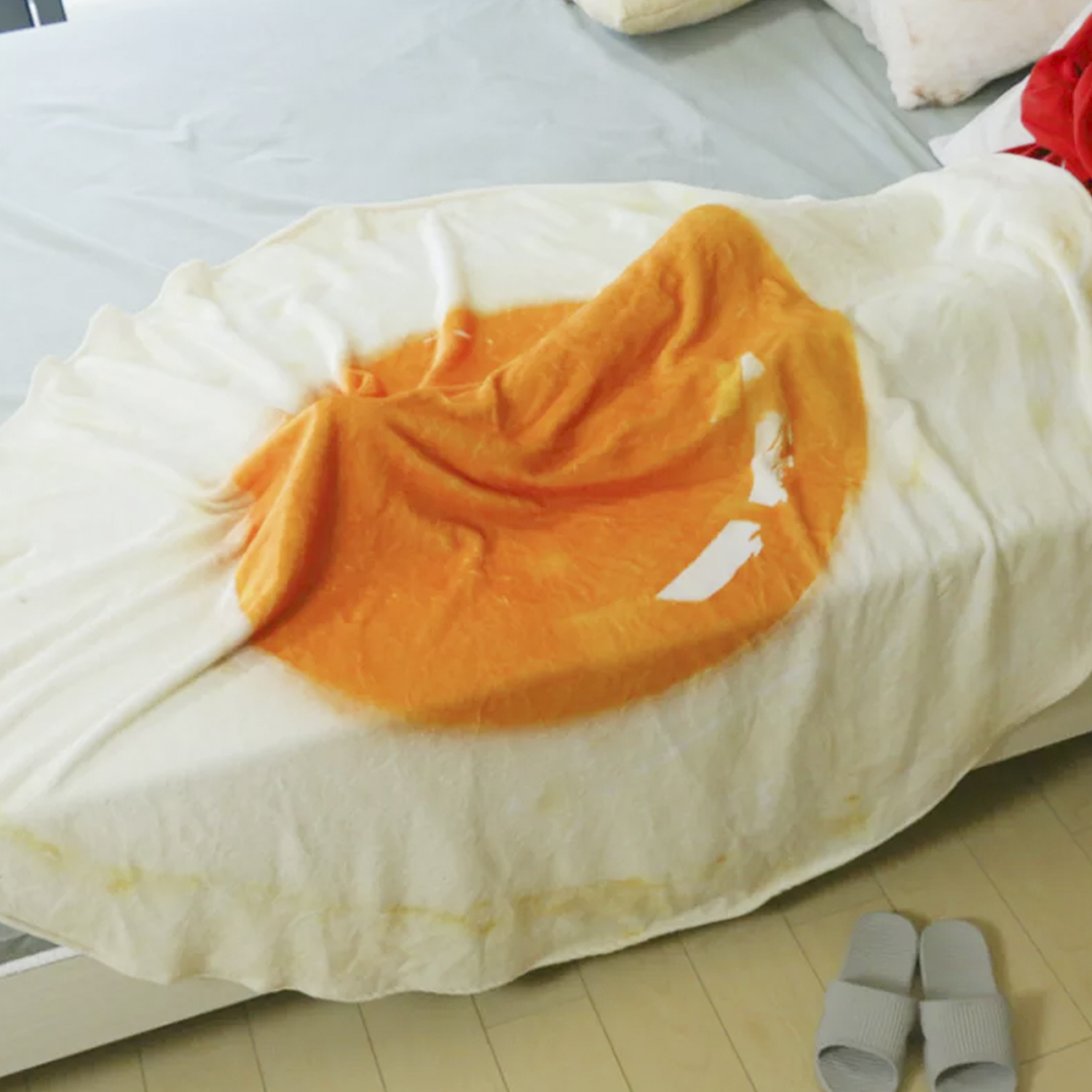 Un letto con una coperta a forma di uovo fritto. Pantofole grigie nelle vicinanze.