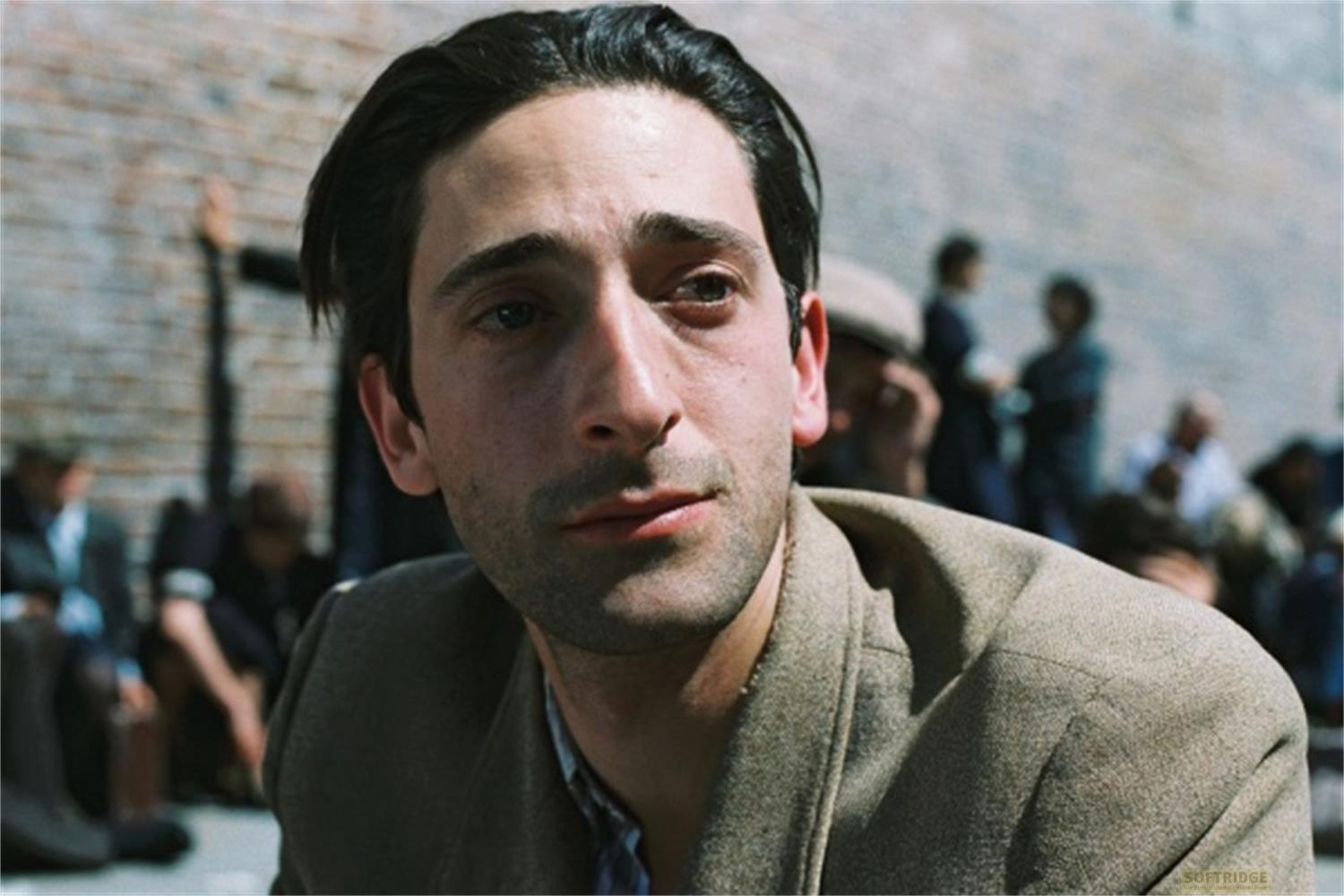 Primo piano di un uomo in giacca beige, che guarda di lato. Persone e muro di mattoni sullo sfondo.