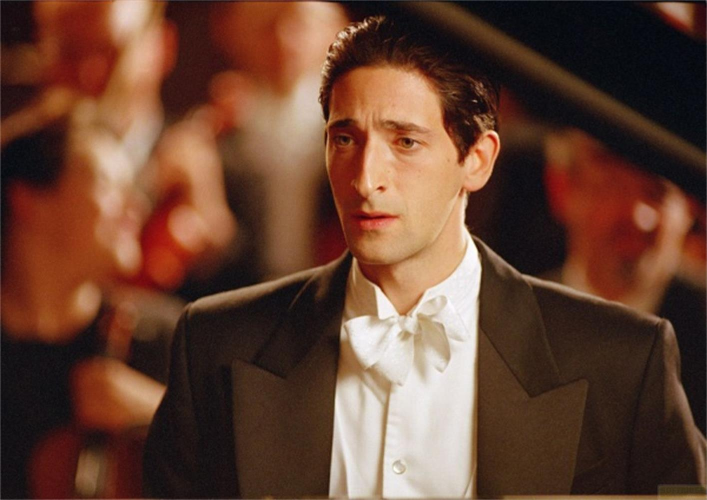 Un uomo in abito nero con papillon, orchestra sullo sfondo.