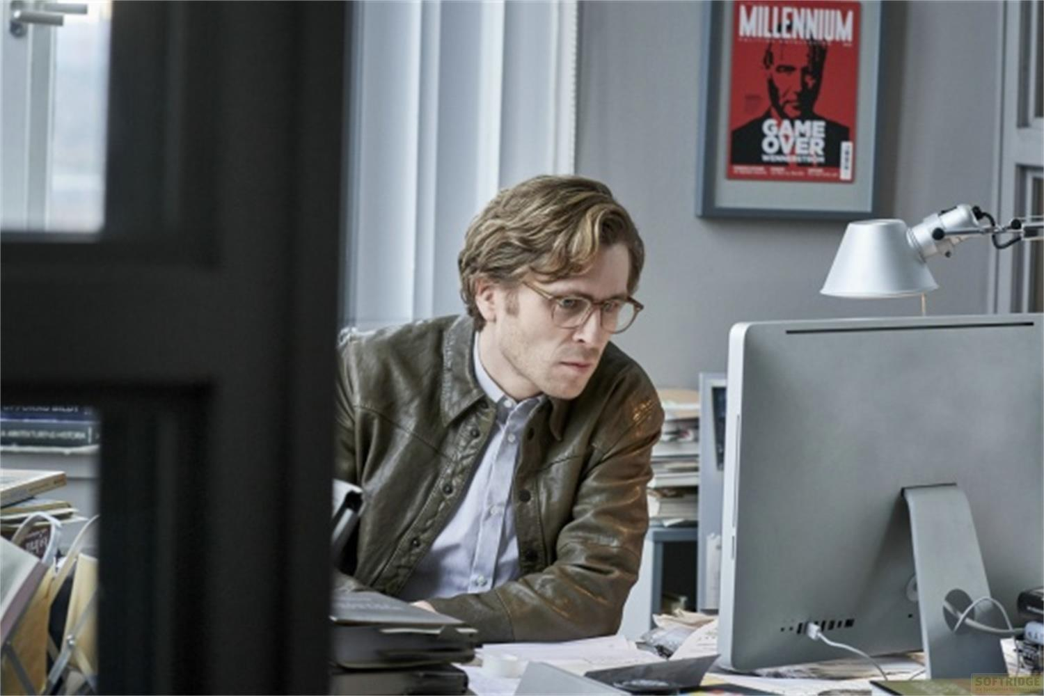 Uomo con occhiali alla scrivania guarda il computer. Poster sul muro.