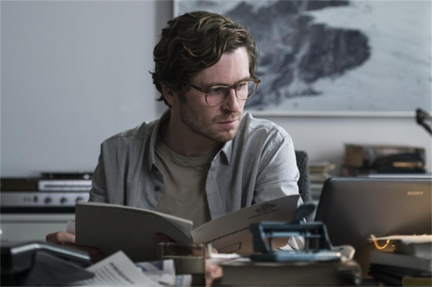 Un uomo con gli occhiali legge documenti alla scrivania. Un laptop è visibile.