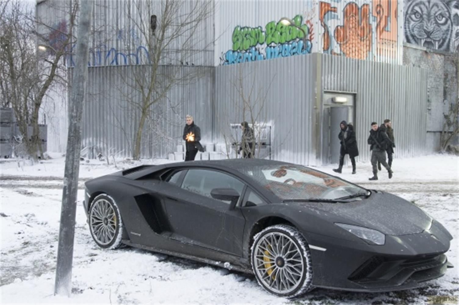 Una Lamborghini nera è nella neve davanti a un muro coperto di graffiti. Si vedono diverse persone.