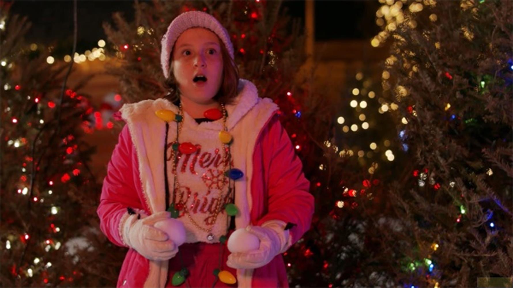 Ragazza con maglione festivo e collana di luci davanti ad albero addobbato.