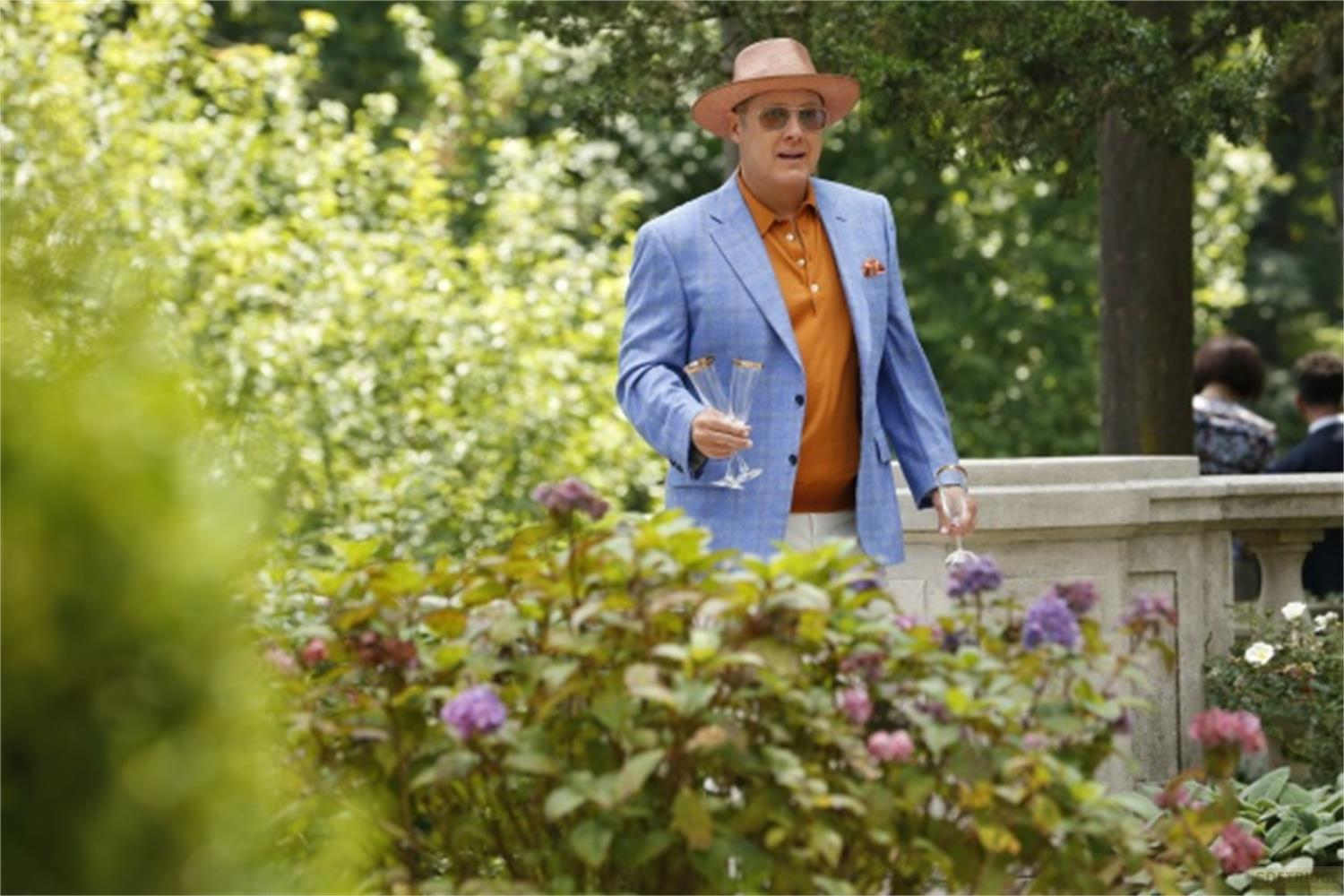 Uomo in giacca blu, camicia arancione e cappello tiene flute di champagne in un giardino.