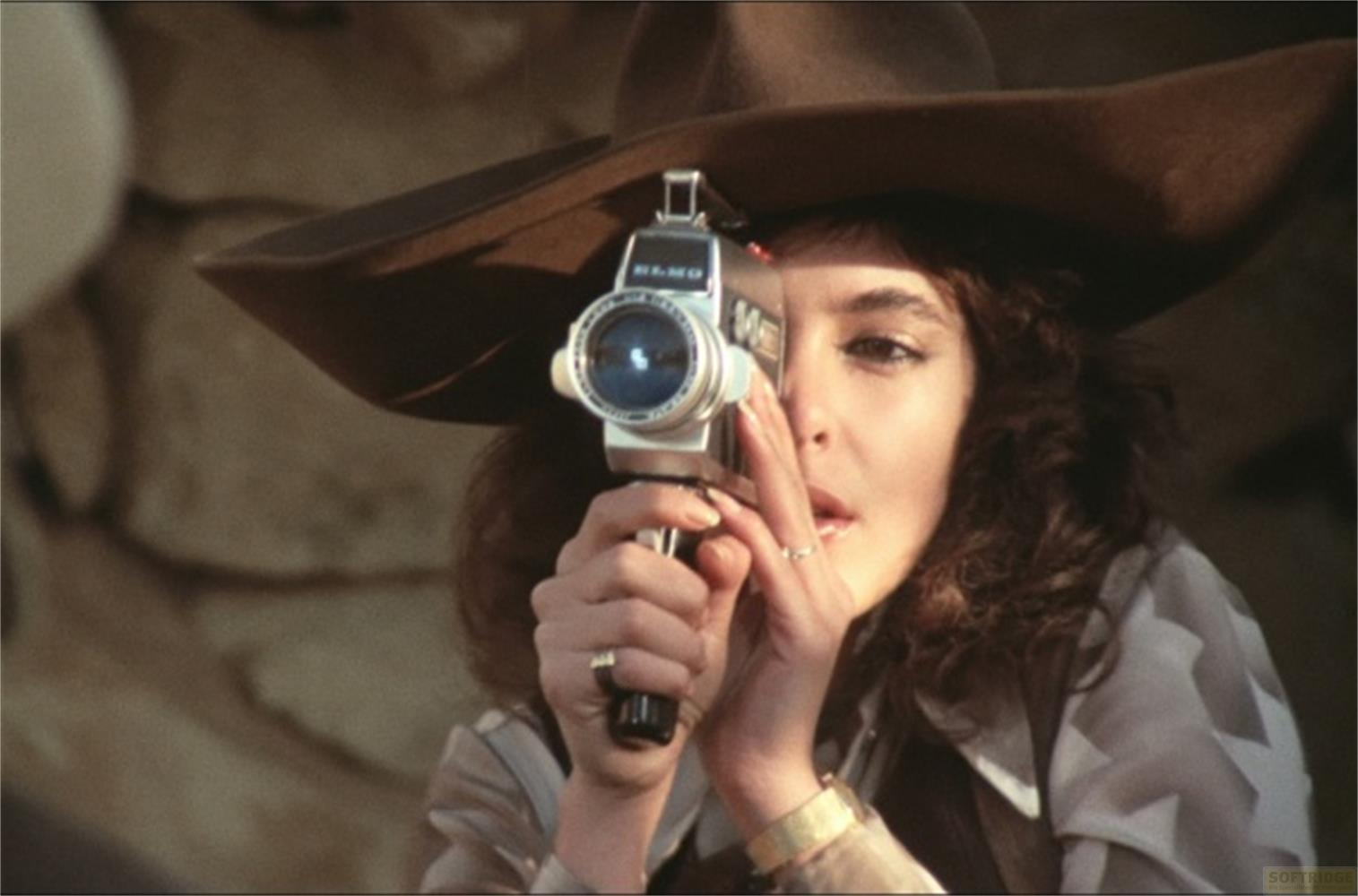 Una donna con cappello filma con una vecchia cinepresa, guardando nell'obiettivo.