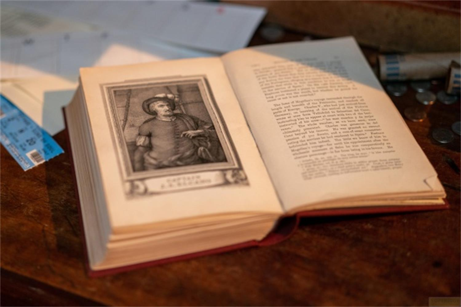 Offenes Buch mit Porträt eines Mannes. Münzen und ein Ticket liegen daneben.