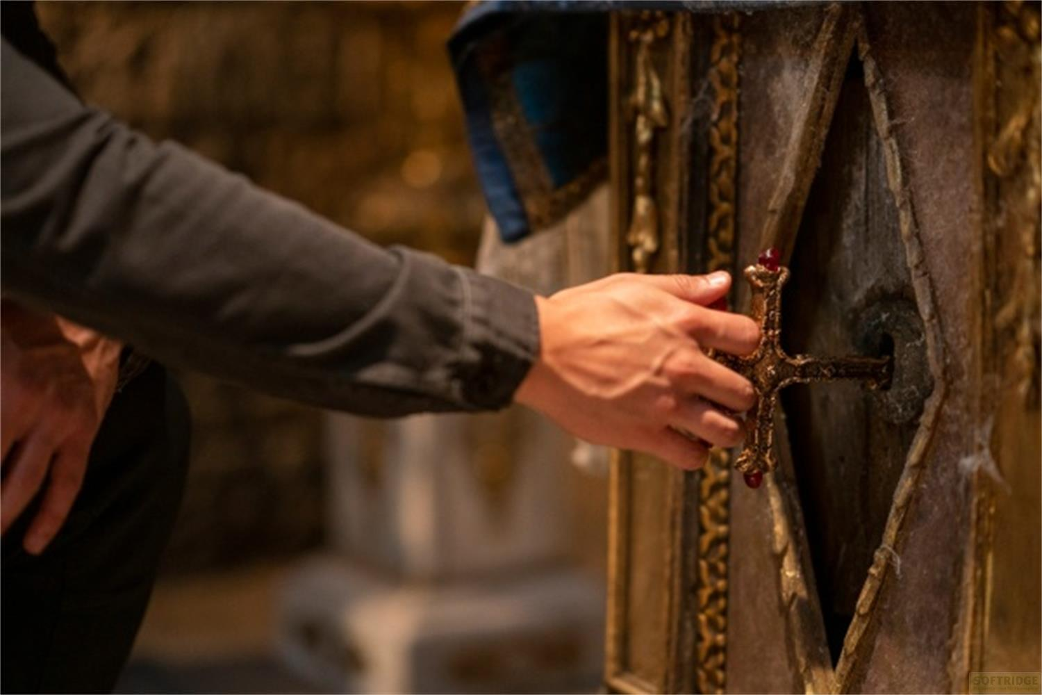 Eine Hand berührt ein goldenes Kreuz mit roten Edelsteinen, das in eine verzierte Holzkiste eingelassen ist.