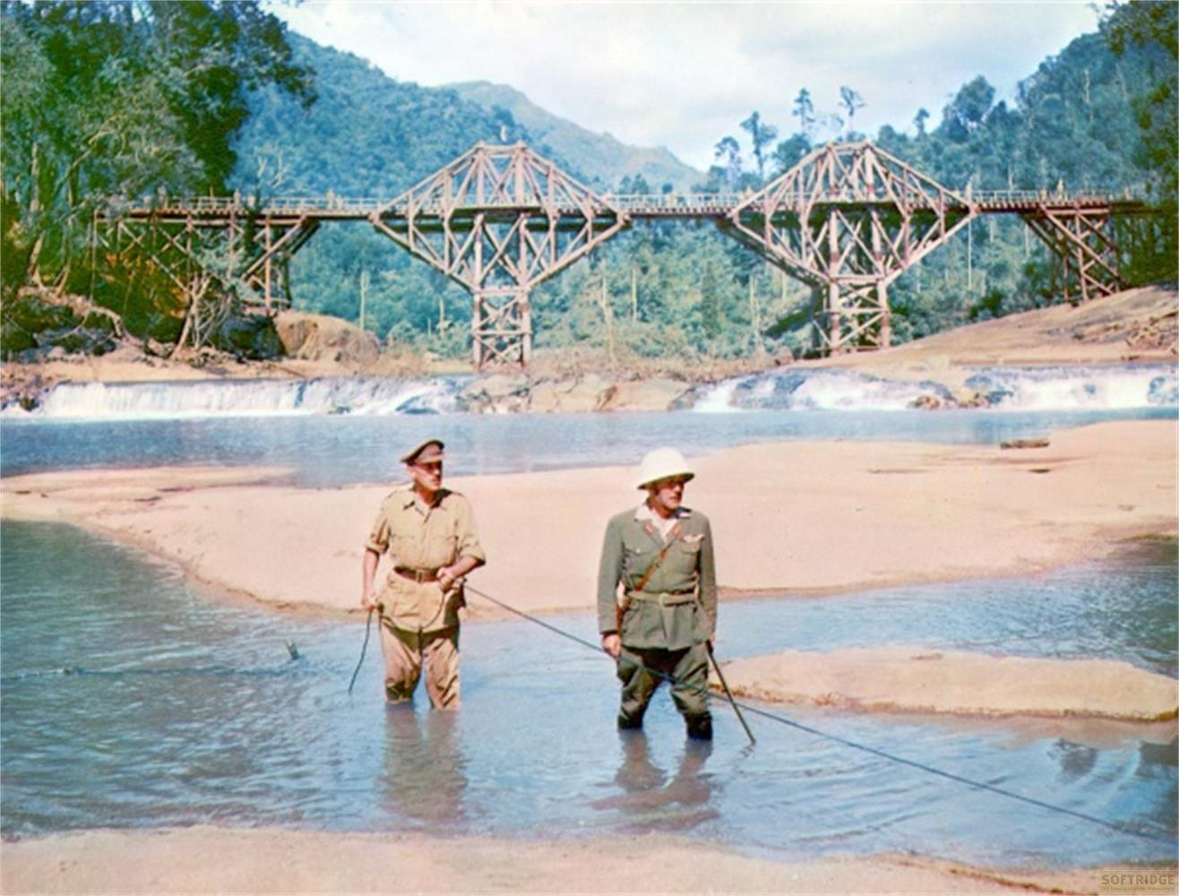 Deux hommes en uniforme se tiennent dans une rivière devant un pont en bois et une cascade.