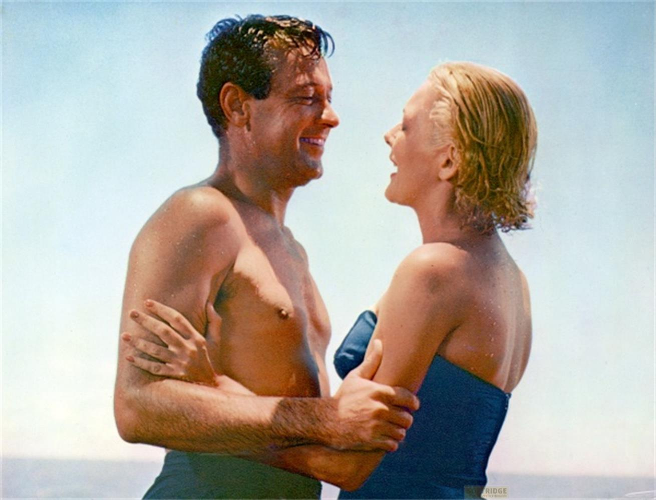 Homme et femme en maillot de bain s'enlaçant devant un ciel bleu.