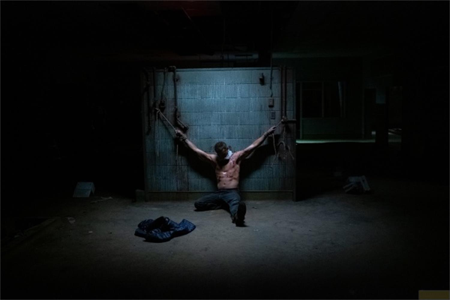 Uomo legato a un muro, in ginocchio, braccia tese, legato con corde.