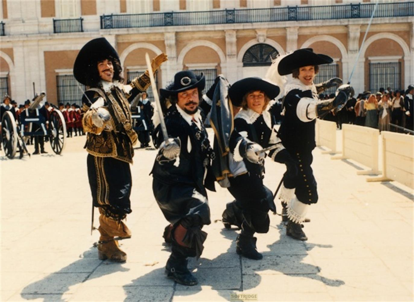 Quattro moschettieri in costume storico con spade davanti a una facciata di edificio.