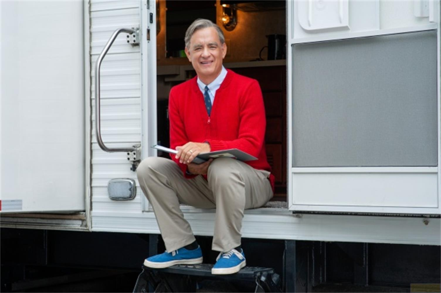 Tom Hanks siede in un cardigan rosso e pantaloni kaki davanti a un camper.