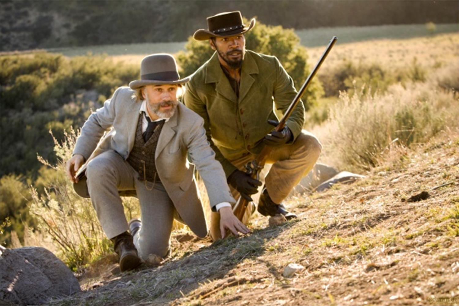 Deux hommes dans le Far West, l'un armé d'un fusil, accroupis sur une colline.