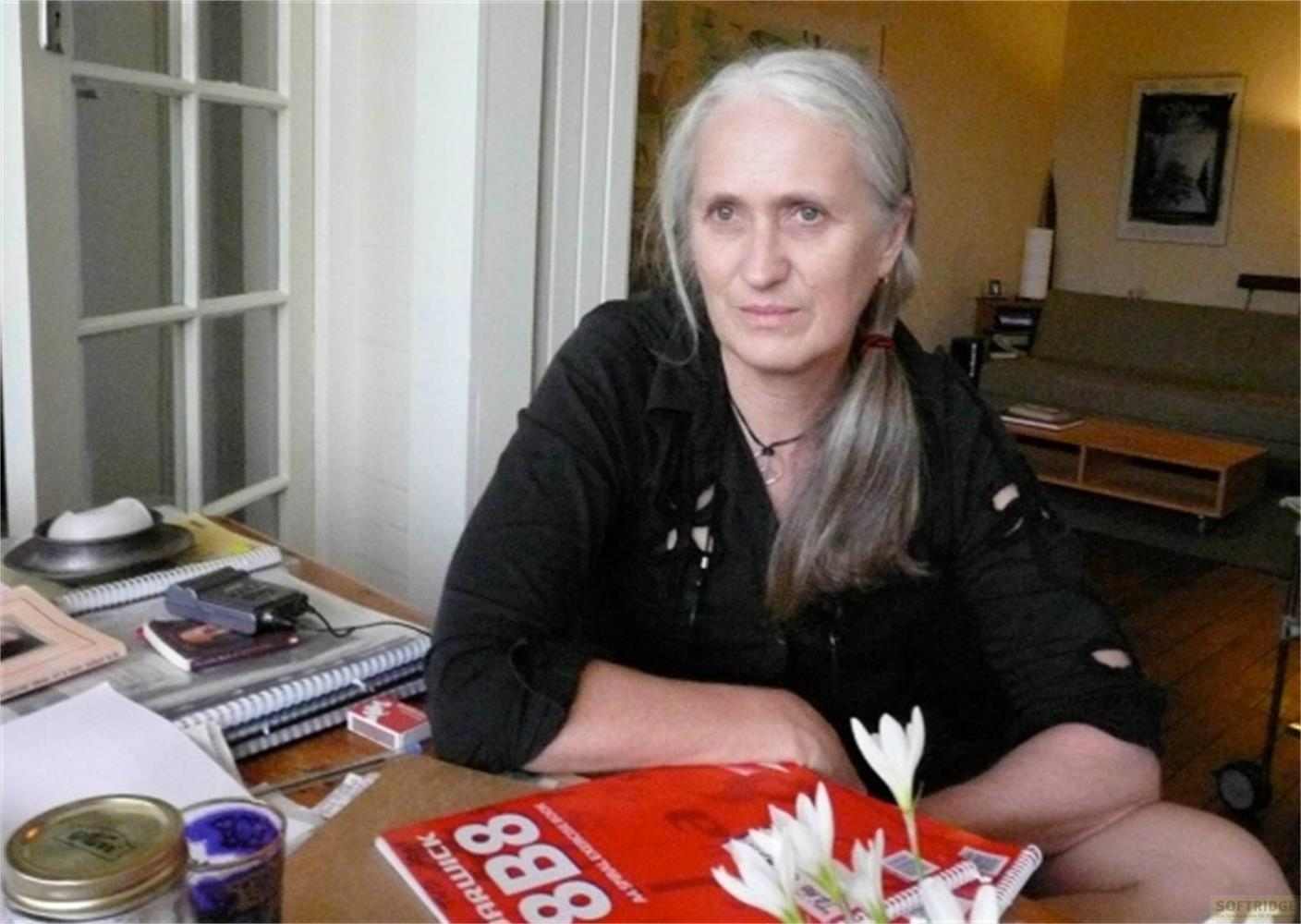 Una donna con lunghi capelli grigi siede a un tavolo con libri e fiori.