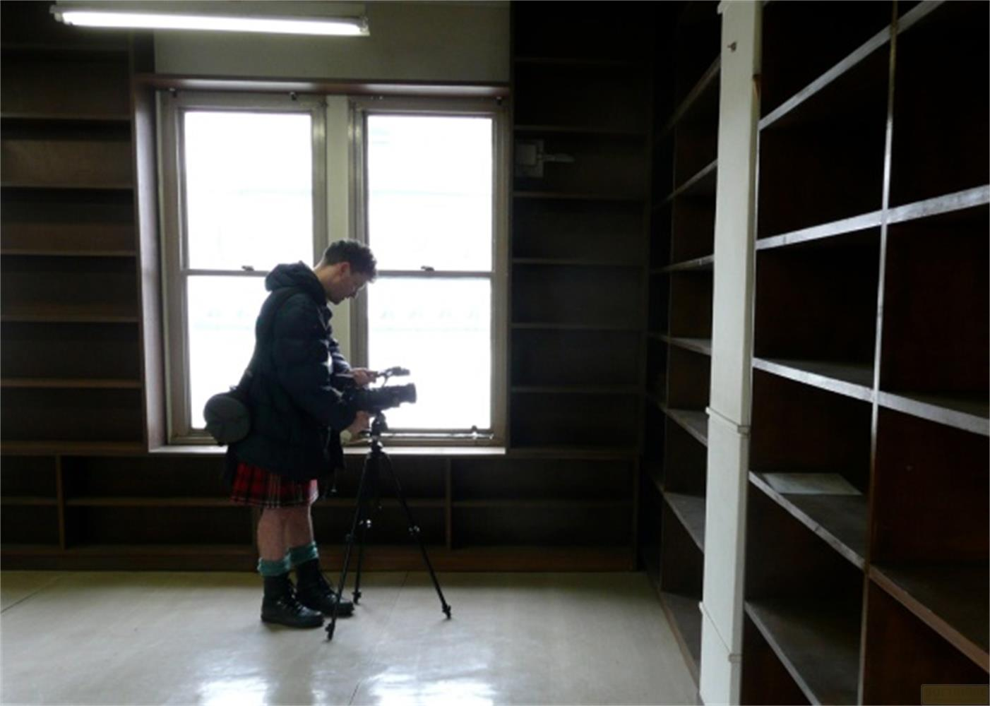 Un uomo in kilt riprende con una telecamera su treppiede in una stanza vuota con scaffali.