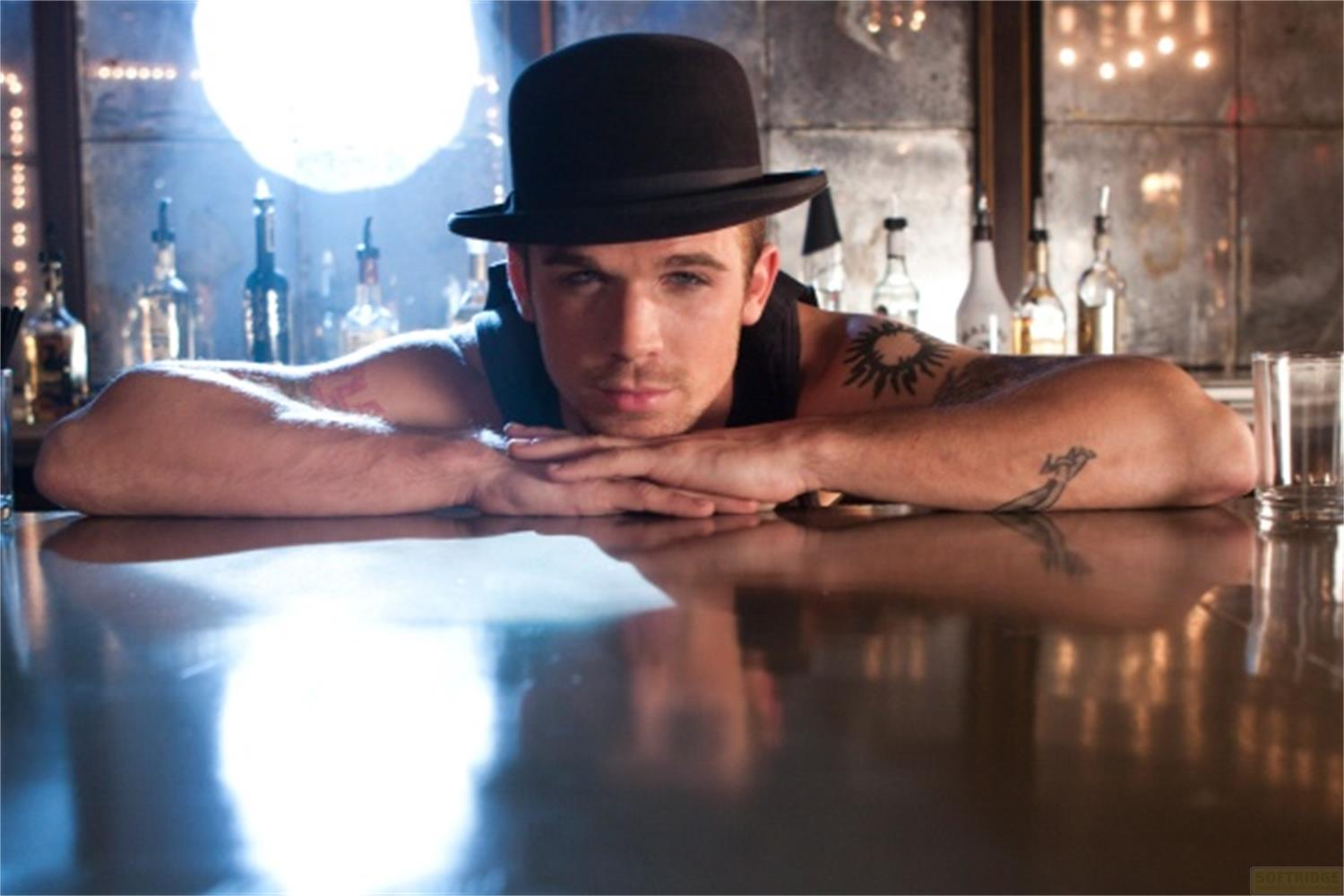 Un homme au chapeau repose sur un bar. Des bouteilles sont visibles au fond.