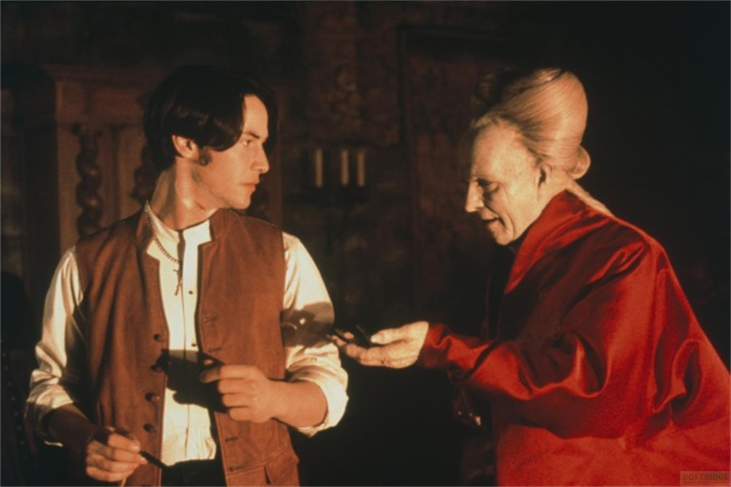 Un homme en gilet et chemise parle à un aîné en robe rouge, tenant de petits objets.