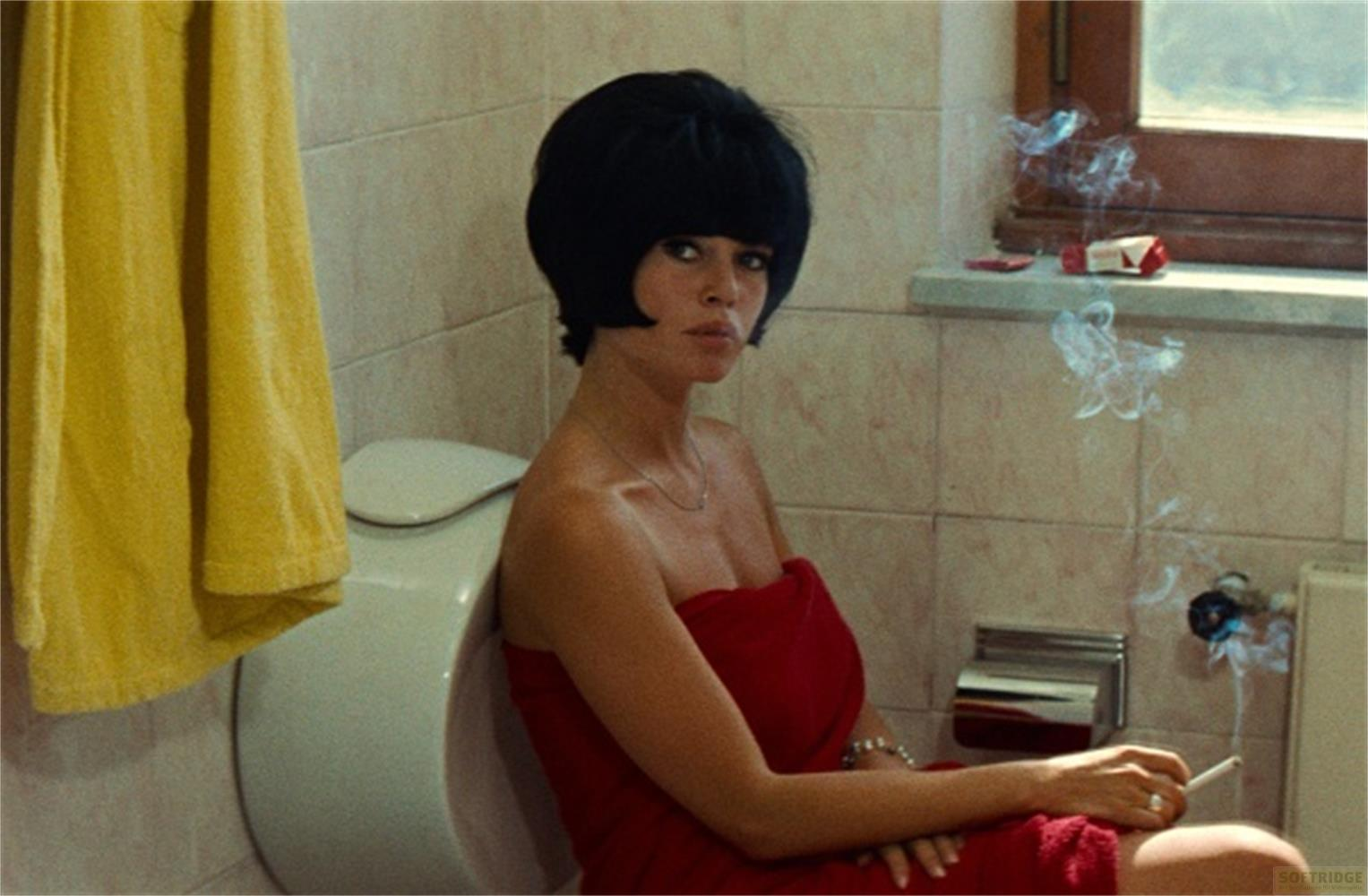 Una donna con capelli neri seduta sul water, avvolta in un asciugamano rosso, fuma.