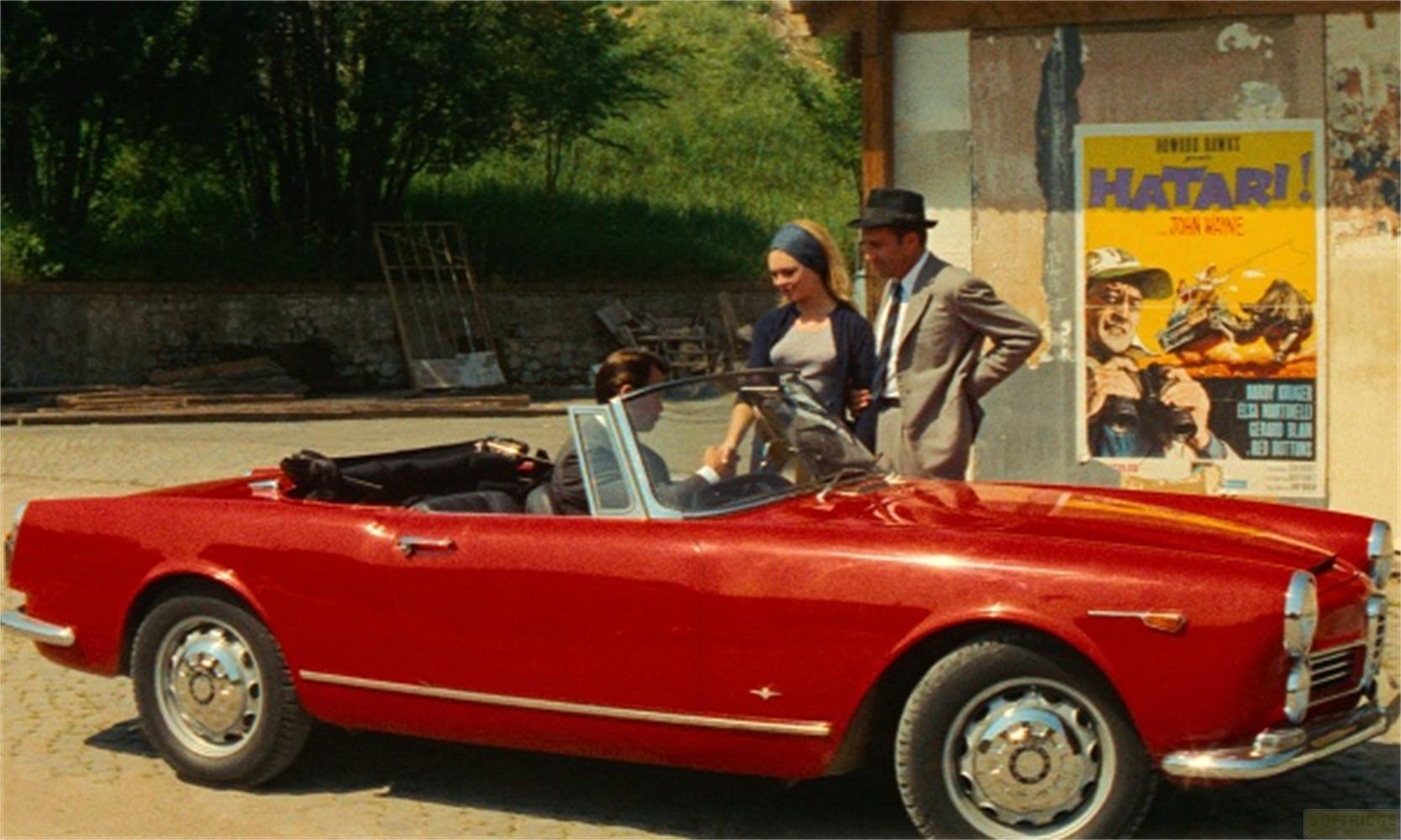 Un uomo e una donna vicino a una cabriolet rossa. Un poster di film sullo sfondo.