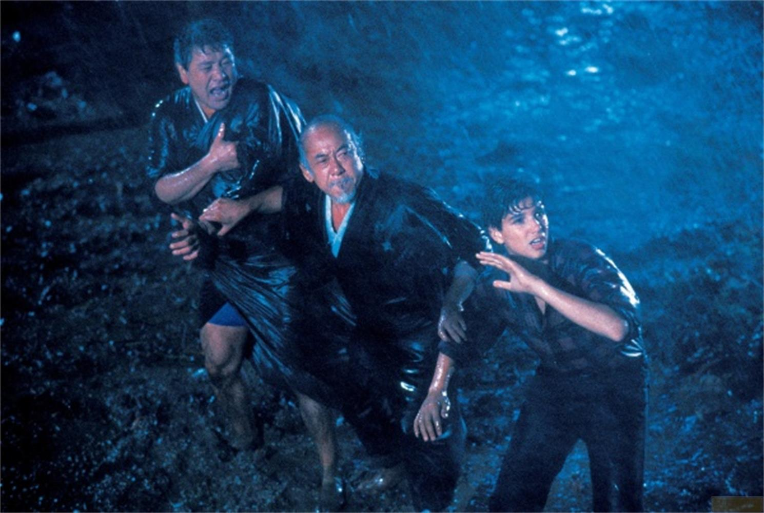 Trois hommes en vêtements sombres courent sous la pluie. L'un est vieux, les deux autres plus jeunes.