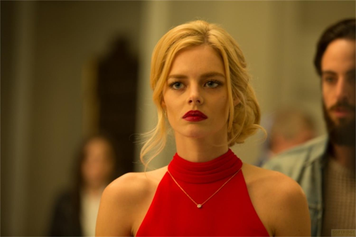 Una donna bionda indossa un vestito rosso con una collana. Figure sfocate sullo sfondo.