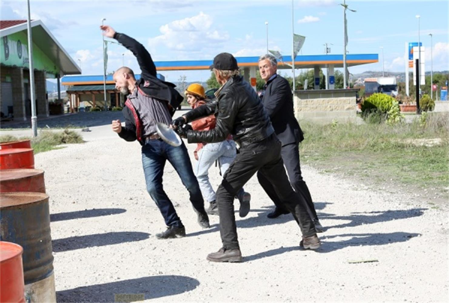 Uomini litigano con una padella davanti a una stazione di servizio e barili rossi.