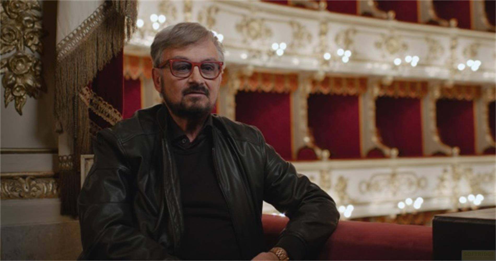 Uomo con occhiali siede in un teatro opulento con tende rosse.