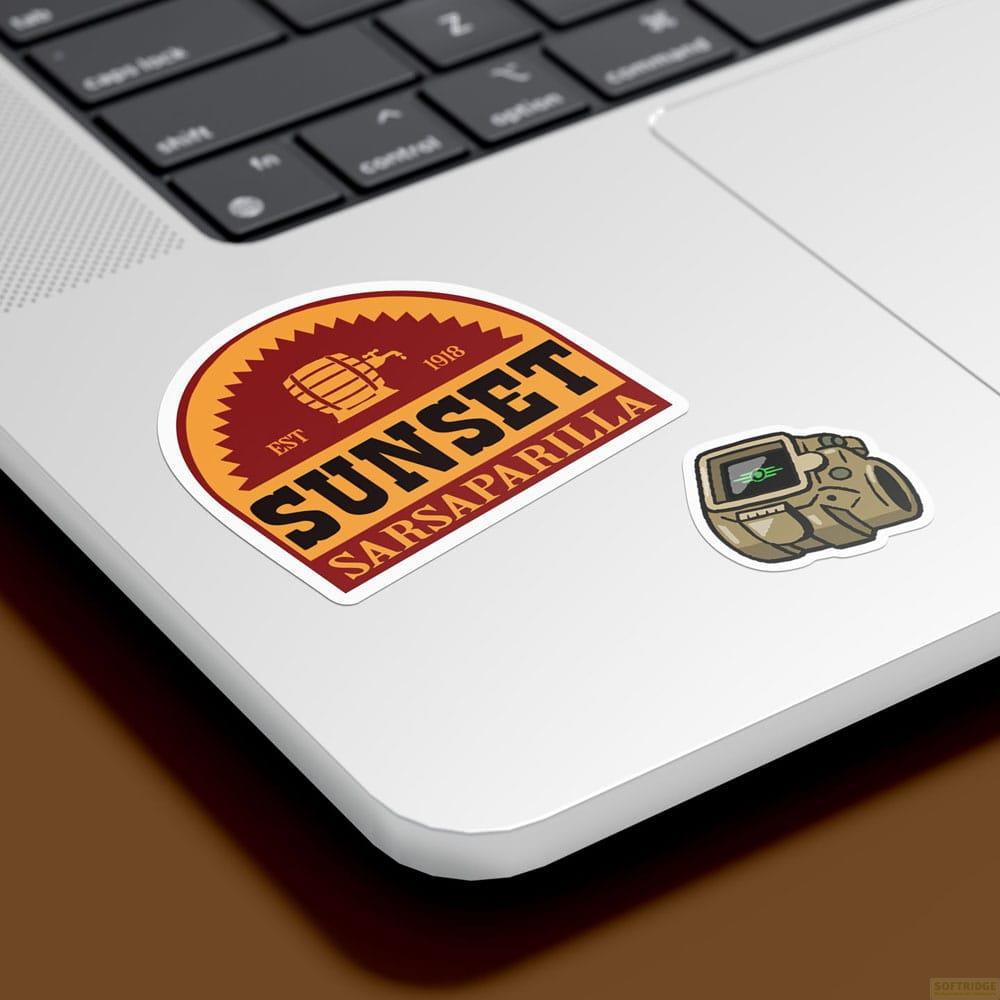 Dos pegatinas en un portátil: un logo redondo de Sunset Sarsaparilla y una mira color canela.