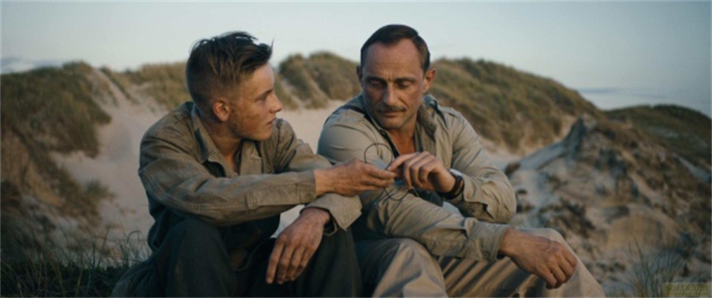 Deux hommes assis dans le sable, entourés d'herbes et de dunes.