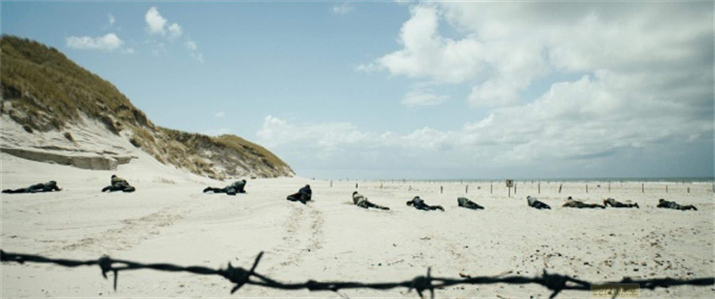 Des soldats sont allongés sur une plage près de dunes, avec du fil barbelé au premier plan.