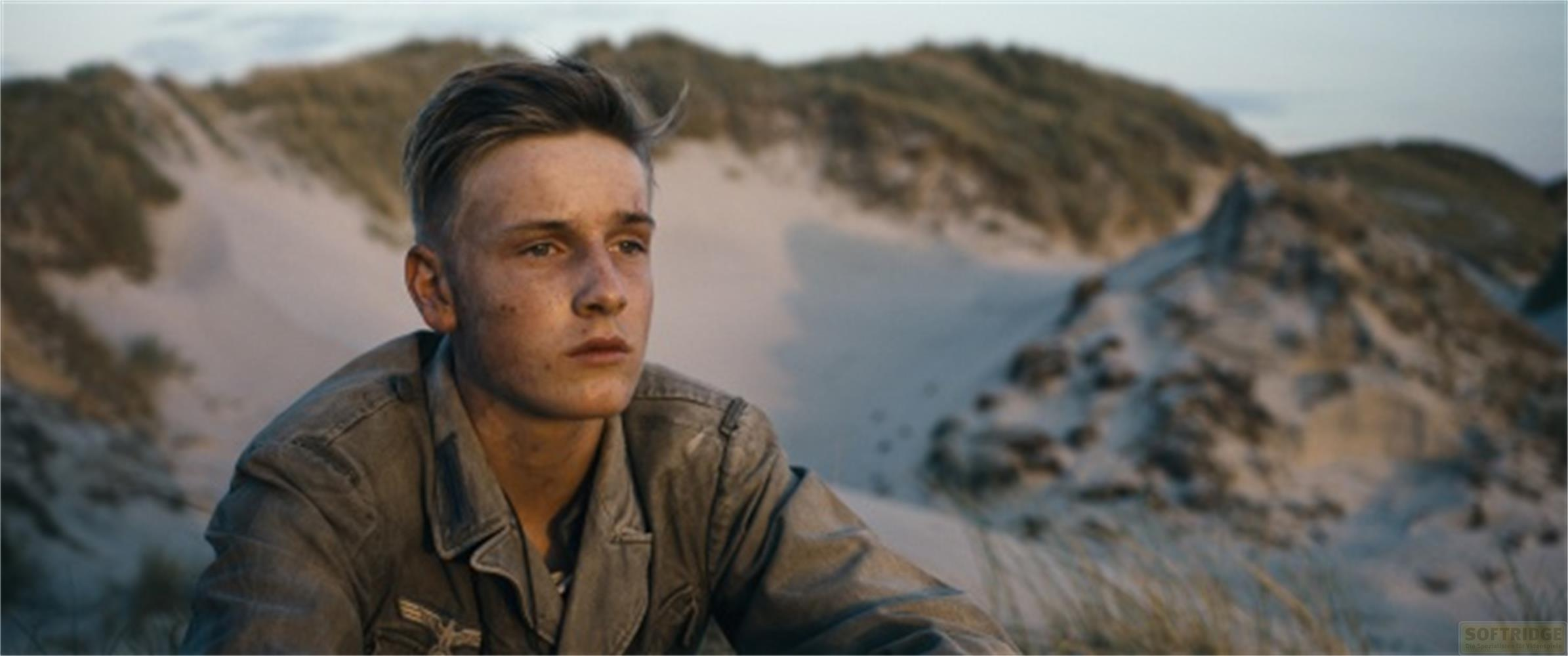 Un jeune homme en uniforme est assis dans le sable, avec des dunes derrière.