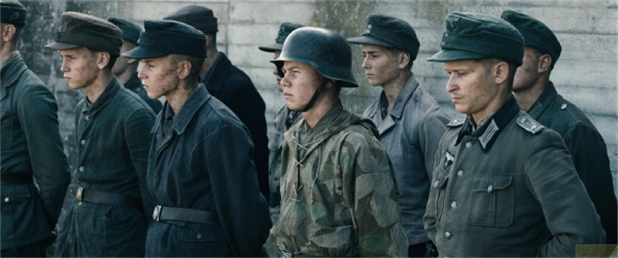 Des soldats en uniforme, certains casqués, forment une ligne devant un mur de béton.