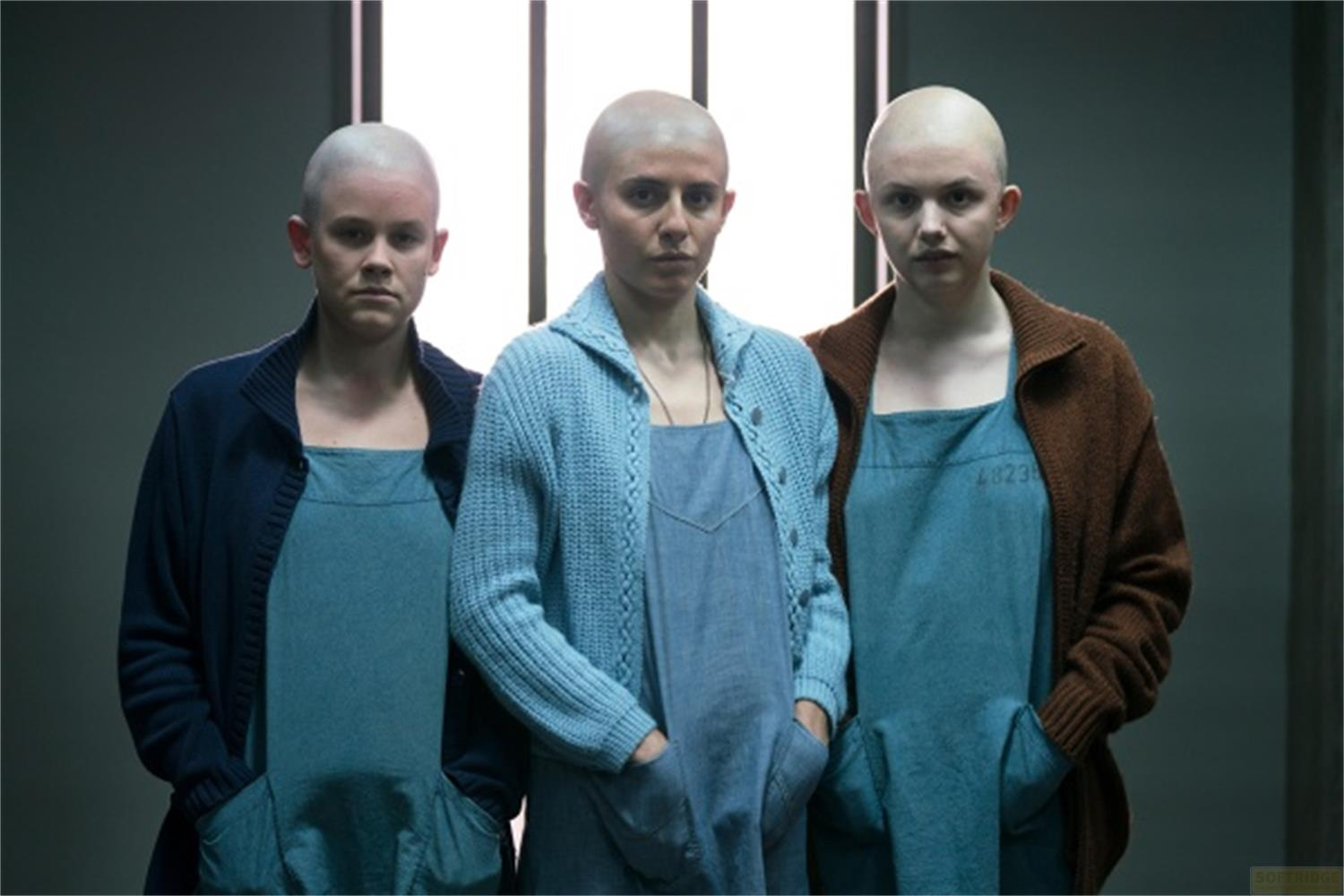 Drei kahlköpfige junge Frauen in blauen Latzkleidern und Strickjacken stehen nebeneinander.