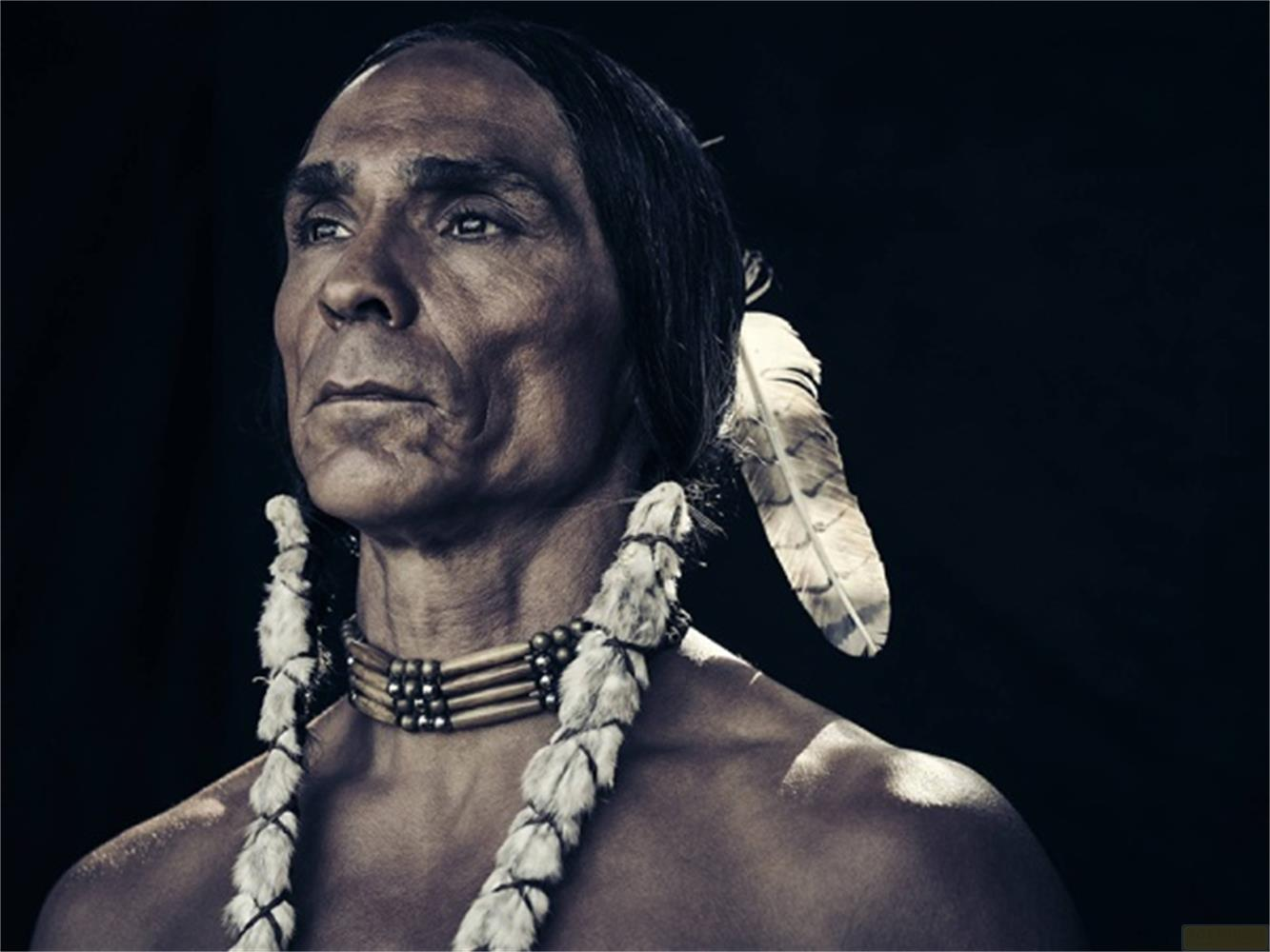Un uomo con lunghi capelli neri, orecchini di piume e una collana d'ossa.