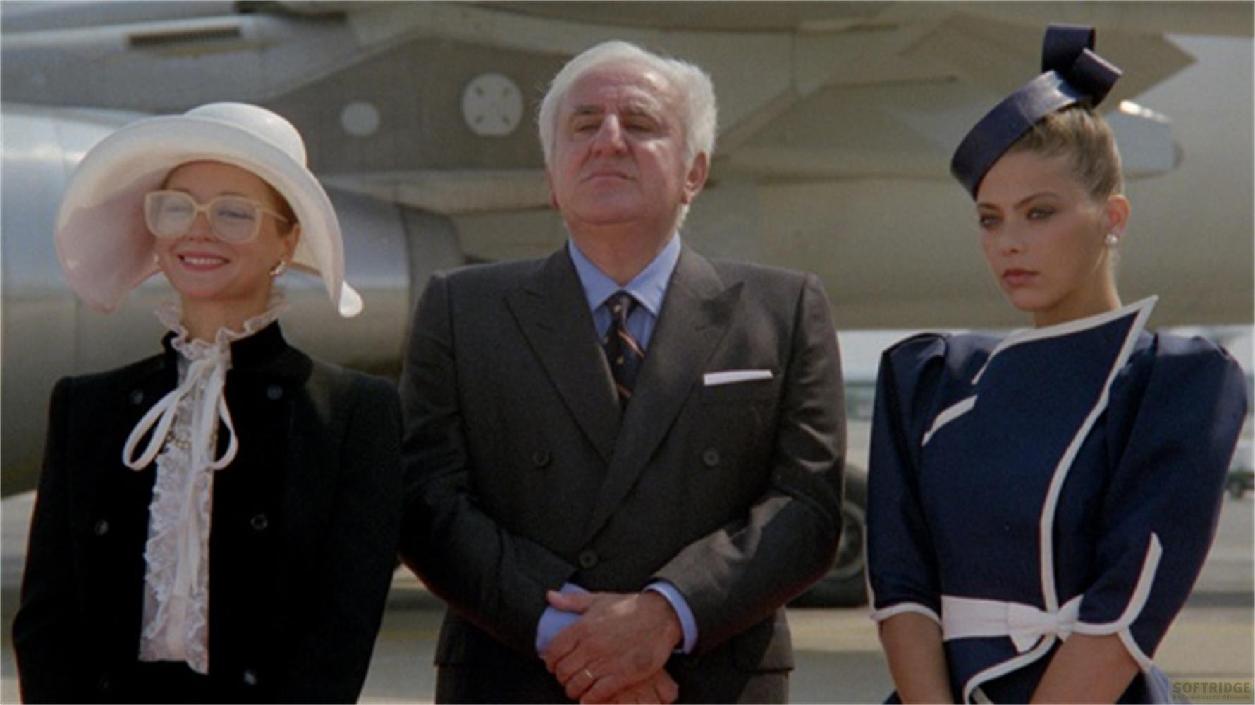 Trois personnes devant un avion : une femme avec un chapeau, un homme en costume, une femme avec un chapeau.