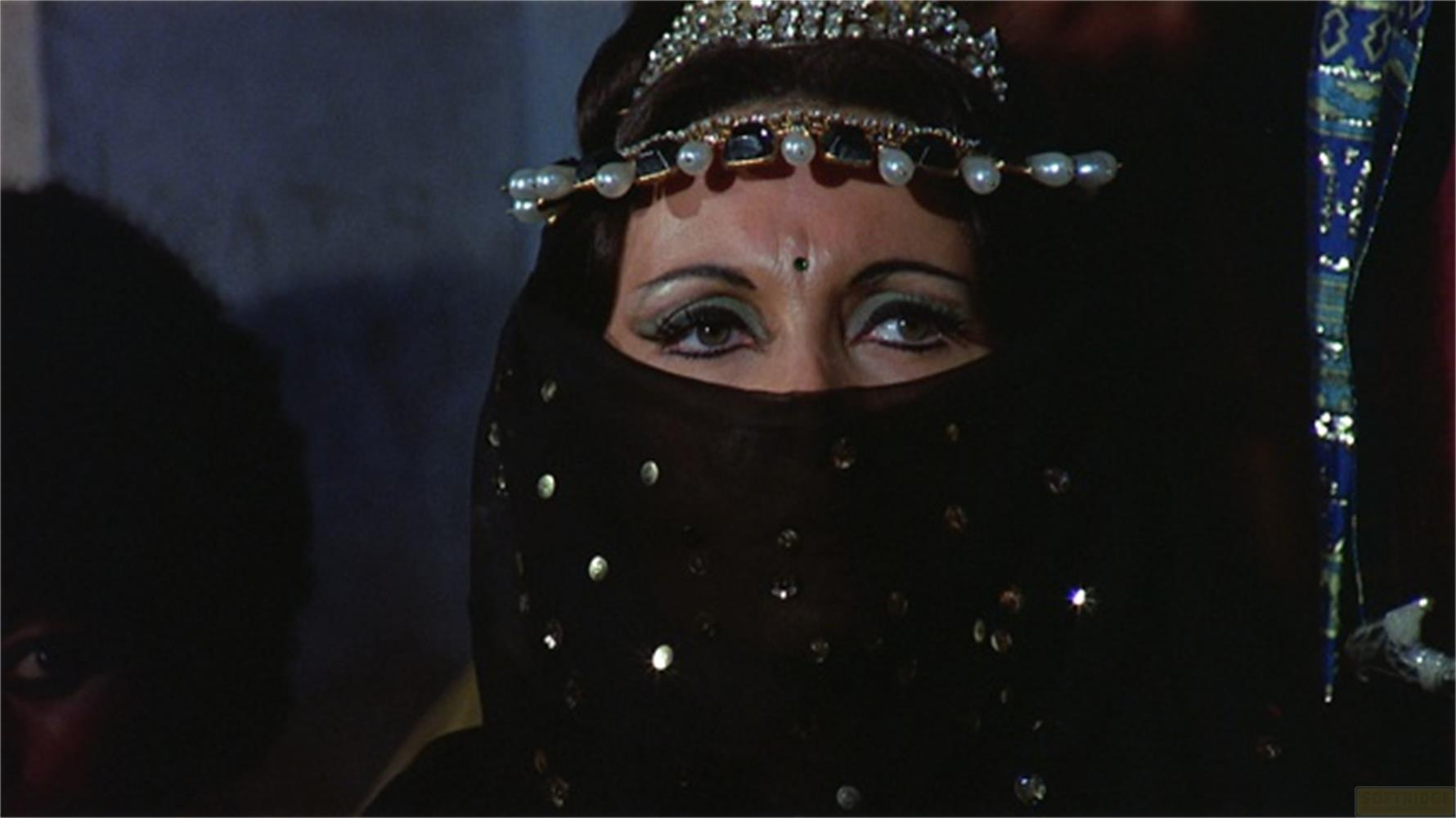 Una donna con velo nero, gioielli e trucco drammatico guarda verso l'alto.